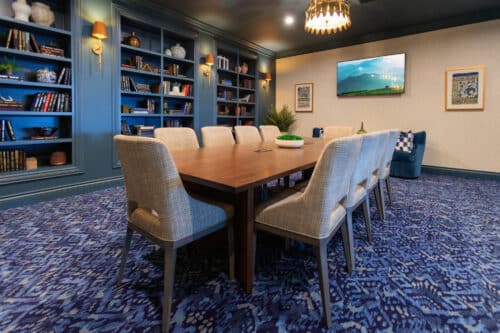 A conference room with a wooden table, eight upholstered chairs, blue patterned carpet, built-in blue bookshelves, and a wall-mounted TV.