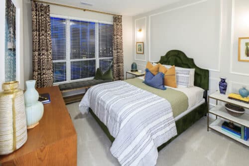 A modern bedroom with a green upholstered bed, striped bedding, decorative pillows, wood dresser, bench by a large window, and patterned curtains.