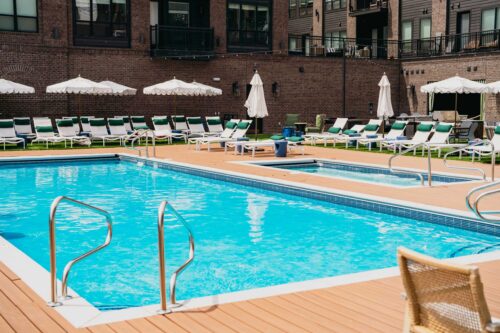 Outdoor swimming pool surrounded by lounge chairs and umbrellas, with a brick building in the background on a sunny day.