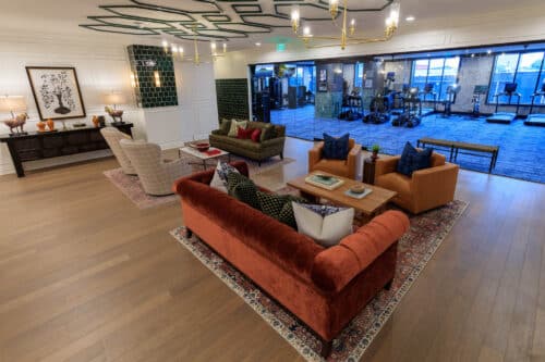 Modern lounge area with velvet sofas, armchairs, and coffee tables on rugs; gym with cardio equipment visible through glass wall in the background.