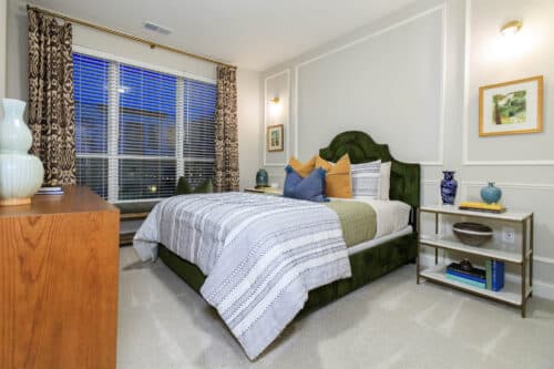 A modern bedroom with a green upholstered bed, patterned bedding, a wooden dresser, two bedside tables with decorative items, and a large window with curtains.
