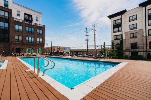 Outdoor swimming pool with wooden deck surrounded by apartment buildings on a sunny day, with lounge chairs and a basketball hoop in the background.