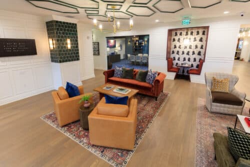 A modern lounge area with tan armchairs, a red sofa, patterned rug, wall art, and a small dining nook in the background, featuring wood flooring and contemporary decor.