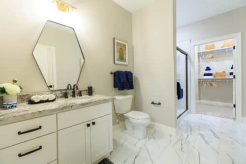Modern bathroom with white cabinetry, granite countertop, hexagonal mirror, toilet, and walk-in shower; closet with shelving and blue towels is visible in the background.