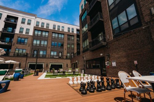 Outdoor courtyard of an apartment complex with a giant chess set, patio seating, green lawn, and multi-story brick and white buildings in the background.