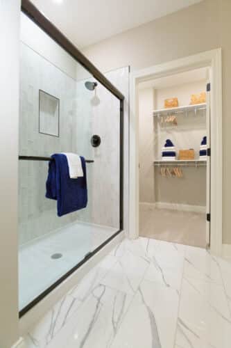 Bathroom with glass shower enclosure, blue and white towels on the door, and a walk-in closet with shelves, baskets, and hanging space in the background.
