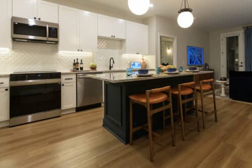 Modern kitchen with white cabinets, stainless steel appliances, a large island with three wooden stools, and a living area with a TV and sofa in the background.