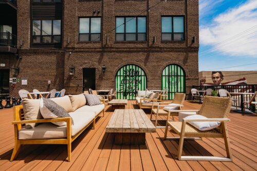 Outdoor rooftop patio with wooden furniture and cushioned seating, set on a deck in front of a brick building with green-striped window arches.