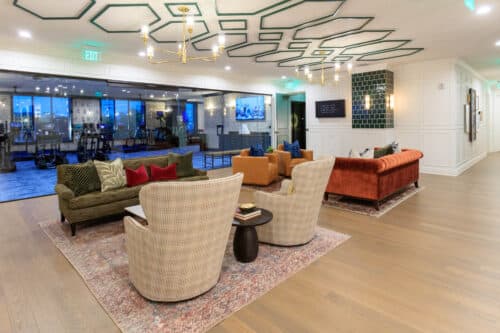 A modern lounge area with upholstered chairs, sofas, accent pillows, and a rug on wood flooring; glass-walled meeting room in the background.