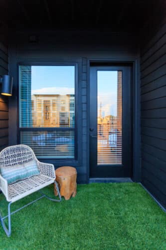Small balcony with artificial grass flooring, a wicker chair, a wooden stump side table, and a glass door and window reflecting nearby apartment buildings.