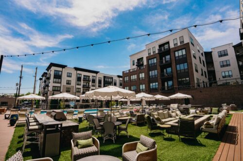 Outdoor lounge area with tables, chairs, and umbrellas on grass, adjacent to a swimming pool and modern apartment buildings under a partly cloudy sky.