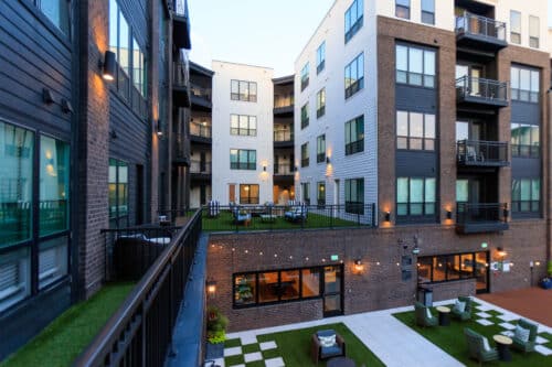 Modern apartment complex with multiple stories, large windows, balconies, and an outdoor courtyard area with seating and grass spaces.