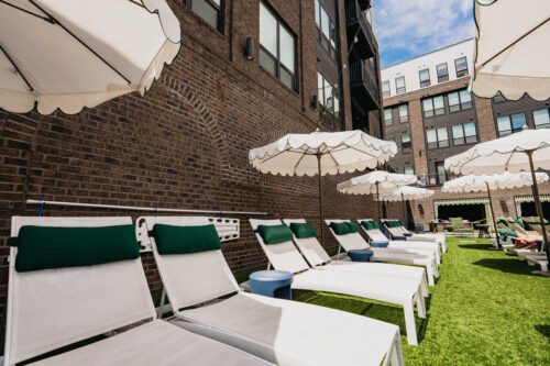 White lounge chairs with green headrests are arranged on artificial grass under white umbrellas beside a brick building, suggesting a modern outdoor relaxation area.