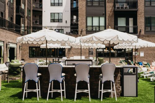 Outdoor bar area with four high chairs, three white umbrellas, and surrounding lounge chairs on artificial grass, set against an apartment building backdrop.