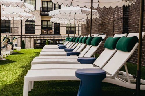 Rows of white lounge chairs with green cushions and blue side tables sit on artificial grass under scalloped white umbrellas beside a brick building.