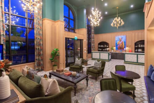 Hotel lobby with high ceilings, large arched windows, green upholstered furniture, patterned curtains, chandelier lighting, and a reception desk with artwork behind it.