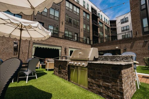 Outdoor communal patio area with a built-in grill, seating, umbrellas, and artificial grass, surrounded by multi-story apartment buildings.