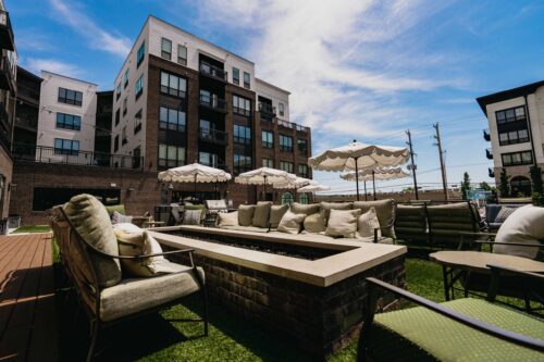 Outdoor patio area with cushioned seating, a rectangular fire pit, and umbrellas, surrounded by modern apartment buildings under a blue sky.