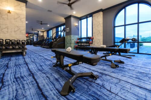 A modern gym with benches, free weights, kettlebells, exercise machines, and large windows letting in natural light.