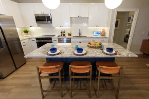 Modern kitchen with white cabinets, stainless steel appliances, and a large island set with three place settings and wooden stools. Fruit basket and decor sit on the countertop.