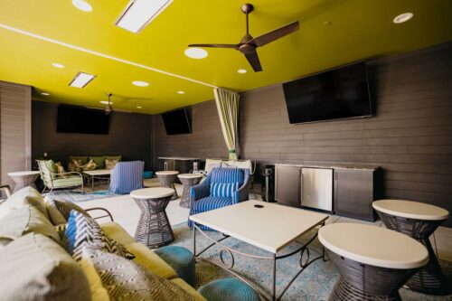 Covered outdoor lounge area with modern furniture, ceiling fans, televisions, a mini fridge, and a yellow ceiling.