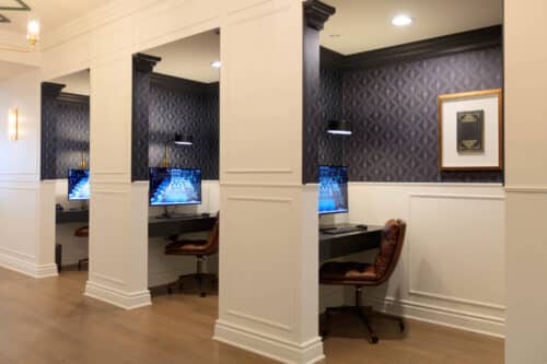 Three individual computer workstations with leather chairs are separated by white partitions in a well-lit room with patterned wallpaper.