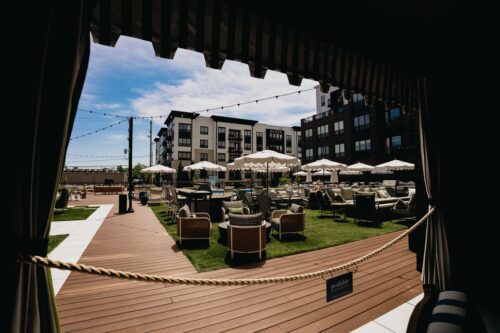 Outdoor patio area with tables, chairs, and umbrellas on artificial grass, surrounded by modern apartment buildings, viewed from behind a roped-off shaded section.