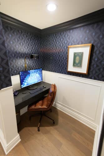 A small home office nook with a desk, computer, brown chair, wall lamp, and framed picture, featuring dark patterned wallpaper and wood flooring.