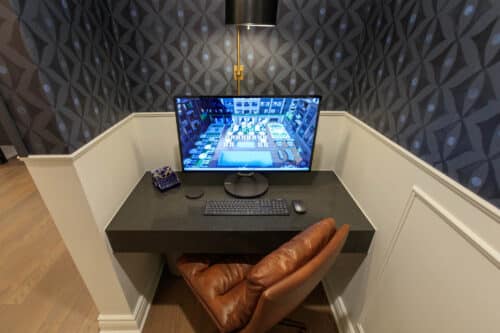 A compact home office with a brown leather chair, a lamp, and a computer displaying a city-building game on a sleek black desk against patterned wallpaper.