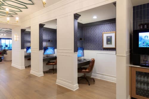 Three computer workstations with chairs are set in individual alcoves along a wall, next to a beverage machine in a modern, well-lit room with wood flooring.
