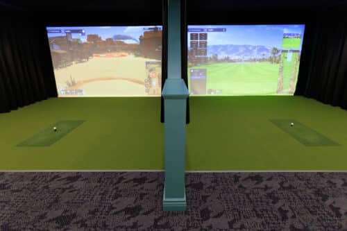 Two indoor golf simulator bays with virtual golf courses displayed on large screens, each bay has a golf ball on artificial turf mats.