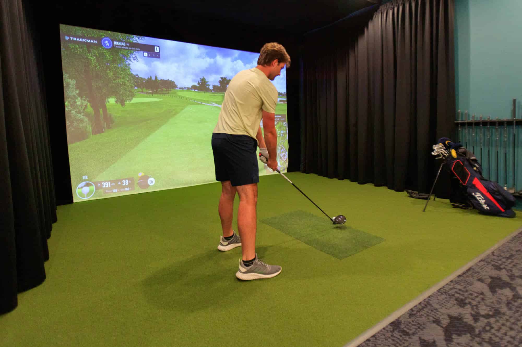 A person prepares to hit a golf ball in an indoor golf simulator, with a virtual golf course displayed on a screen in front of them.