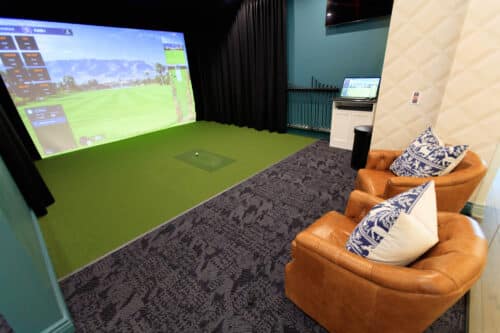 Indoor golf simulator setup with a large screen, golf mat, two brown leather chairs with patterned cushions, and a small monitor on a stand.