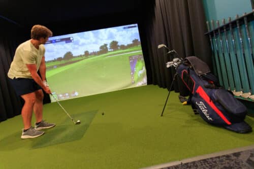 A man prepares to hit a golf ball in an indoor golf simulator room, with a golf bag and clubs nearby and a projected course on the screen.