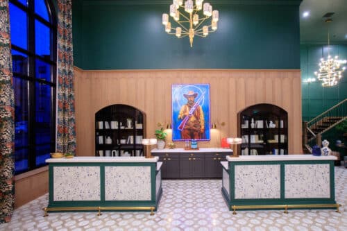 Hotel reception area with two front desks, bookshelves, a large portrait of a cowboy on the back wall, chandeliers, and patterned flooring.