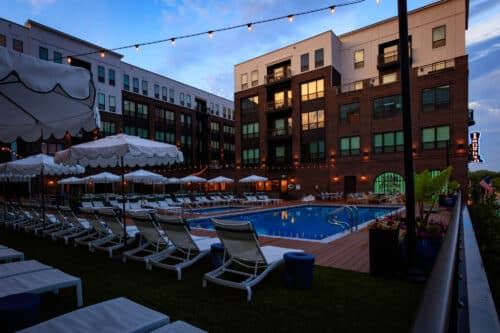 Outdoor pool area at an apartment complex with lounge chairs, umbrellas, string lights, and multi-story buildings in the background at dusk.