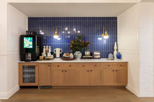 A coffee and snack station with a drink machine, cups, pastries, and decorative vases on a wooden counter, set against a blue tiled backsplash.