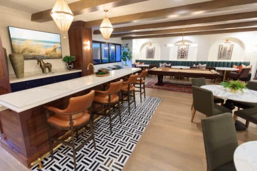 A modern kitchen and dining area with a marble island, leather barstools, geometric rug, lounge seating, wall art, and large pendant lights.