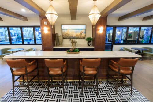 A modern lounge area with a marble island, four brown leather chairs, pendant lights, and wall art, with large windows in the background.