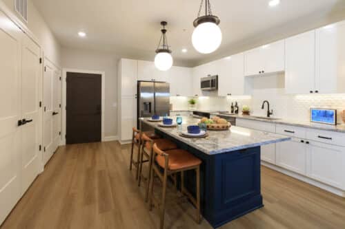 Modern kitchen with white cabinets, stainless steel appliances, a granite island with blue base, three bar stools, pendant lights, and wood flooring.