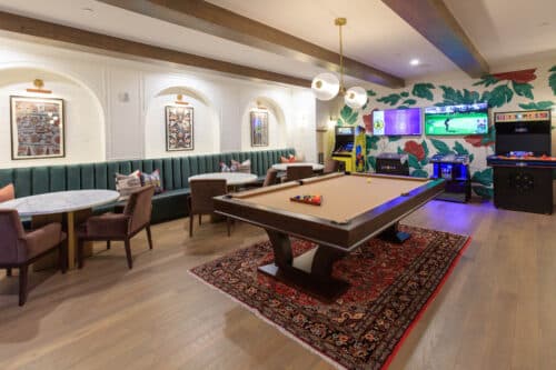A game room with a pool table on a rug, booths with tables and chairs, wall art, an arcade machine, and a TV displaying sports in the background.