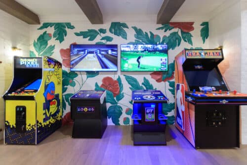 Four arcade machines and two wall-mounted screens displaying sports games are set up against a floral mural in a game room with wood flooring and exposed ceiling beams.