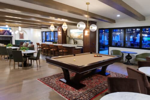 A modern lounge with a pool table on a rug, dining tables, bar seating, overhead lights, and large windows showing an outdoor patio.