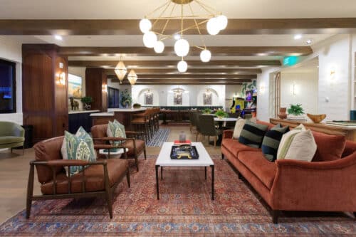 A modern lounge with armchairs, sofas, a coffee table, patterned rug, and overhead globe lights, featuring exposed ceiling beams and decorative plants.