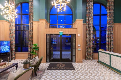 Hotel lobby with large arched windows, patterned tile floor, chandeliers, floral curtains, a reception desk, and a rug labeled "Langham Boutique Collection.