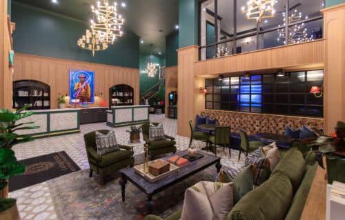 Stylish hotel lobby with green walls, modern chandeliers, velvet chairs, a front desk, wall art, and a mix of seating areas under warm lighting.