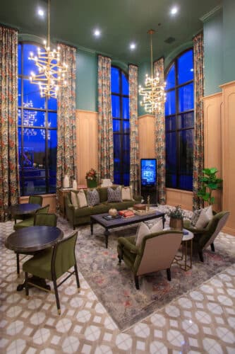 A spacious lounge with high ceilings, patterned floor tiles, tall floral curtains, modern chandeliers, green armchairs, and a large rug around a central coffee table.
