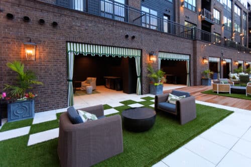 Outdoor seating area with wicker chairs, a round table, artificial grass, potted plants, and open cabanas along a brick building facade.
