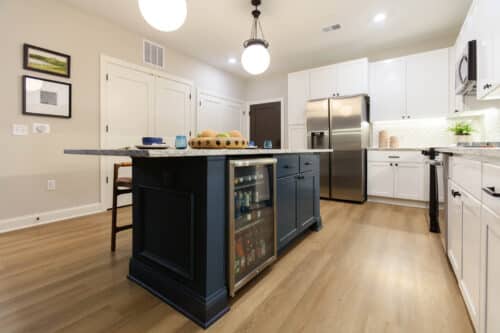 Modern kitchen with white cabinets, stainless steel appliances, and a navy island featuring built-in shelving and a glass-front mini fridge, set on light wood flooring.