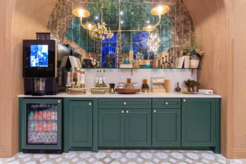 A refreshment station with green cabinets, a beverage cooler, a coffee machine, snacks, and condiments, set against a mirrored wall with gold lighting fixtures above.